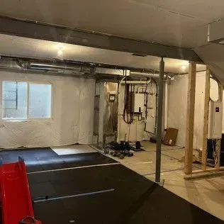 Basement with exposed pipes, framing, and a utility room. Black rubber flooring in foreground.