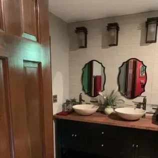 Bathroom with two sinks, two mirrors, sconces, and a dark wooden cabinet, with a partially open wooden door.