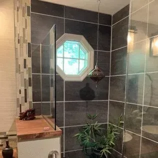 Bathroom with black tile shower, octagon window, glass shower divider, and potted plant.