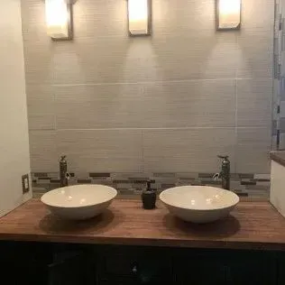 Two bowl sinks on a wooden countertop with faucets and a soap dispenser, tile backsplash.
