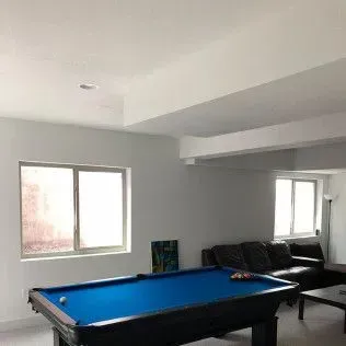 A pool table with a blue felt top in a room with windows and a dark leather couch.