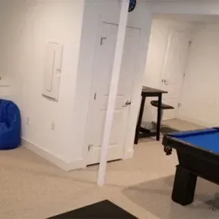 A white cat jumps on a black mat in a basement with a pool table, bar stool, and bean bag chair.