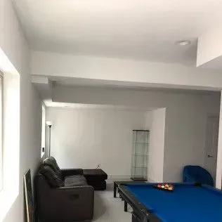 A spacious, well-lit basement rec room with a pool table, dark leather sofa, and a glass display cabinet.