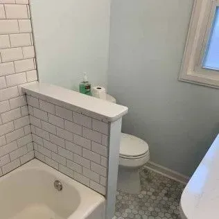 Bathroom with a white bathtub and toilet, separated by a tiled wall. Light blue walls and a small window are visible.