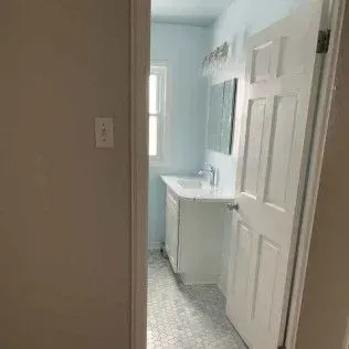 Bathroom interior with sink, mirror, white door, window. Light blue walls, gray tile floor.