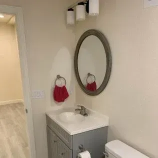 Small bathroom with an oval mirror, sink, and toilet; gray cabinet; white walls.