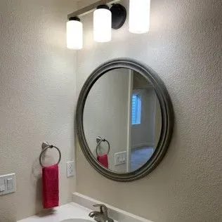 Bathroom with round mirror, gray frame, above a sink. Pink towel hangs from a silver ring. Three-light fixture.