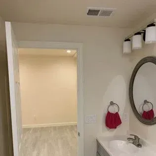 Bathroom interior, open door to empty room, vanity with sink, mirror, towel holder, overhead lights, neutral colors.