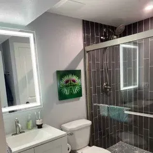 Bathroom with gray walls and a dark tiled shower. Illuminated mirror, white vanity, and toilet. Art on wall.