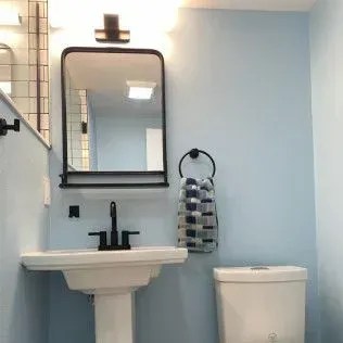 Bathroom with a square mirror, pedestal sink, towel ring, and toilet on a light blue wall.