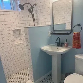A bathroom with a tiled shower, pedestal sink, and a black-framed mirror over the sink. The walls are light blue.