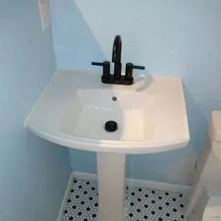 White pedestal sink with black faucet against a light blue wall, tiled floor.