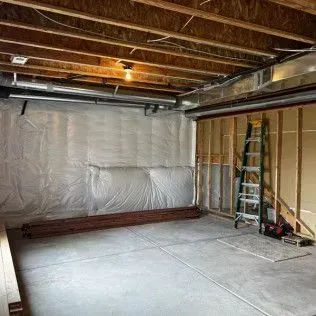 Unfinished basement with concrete floor, wood framing, insulation, and exposed ceiling beams. A ladder leans against a wall.