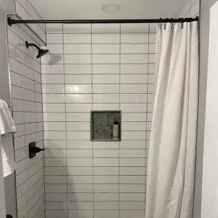 White-tiled shower with black fixtures and a white curtain. A small built-in shelf holds a bottle.