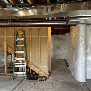 Basement under construction; unfinished wall with ladder, ductwork overhead, insulation rolls to the right.