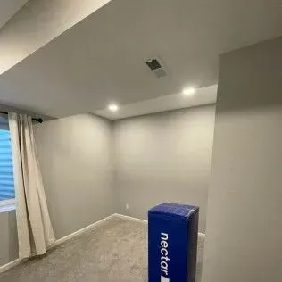 A small, empty room with grey walls and carpet, a window with curtains, and a Nectar mattress box.