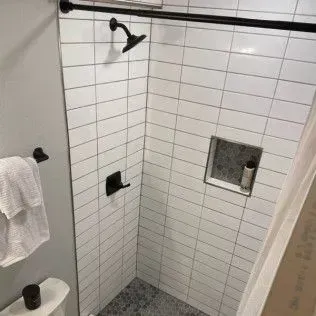 Small white-tiled shower with black fixtures; a towel rack and toilet are visible on the left.