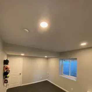 Basement room with white walls, a window with a blue light, and a door on the left.
