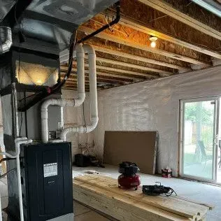 Basement under construction, with furnace, pipes, plywood, and a sliding glass door.