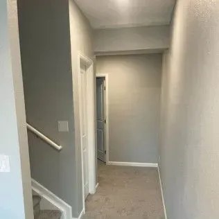 Narrow hallway with gray walls, light carpet, and a door at the end. Staircase visible on the left.