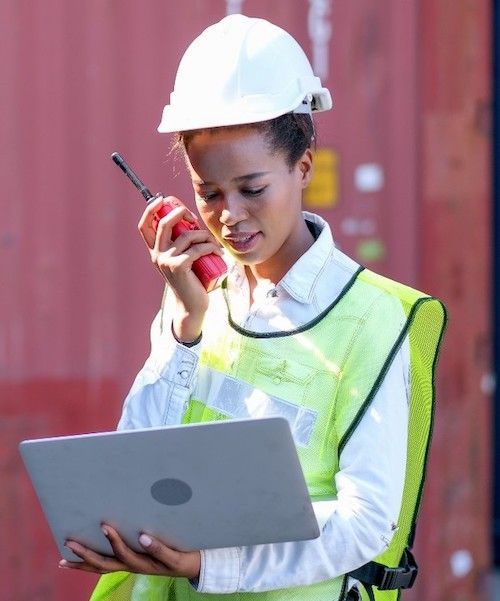 A woman wearing a hard hat and safety vest is holding a laptop and talking on a walkie talkie.