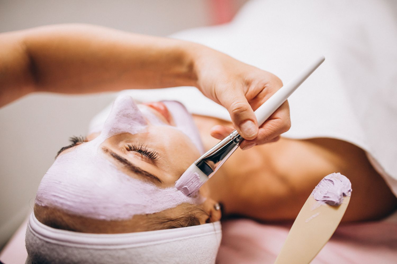 Person receiving a facial mask application in a spa; brush applying purple mask to face.