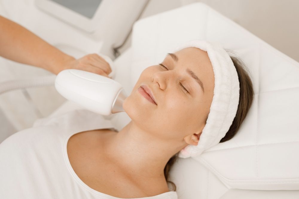 Woman receiving facial treatment with a handheld device, lying on a white bed.