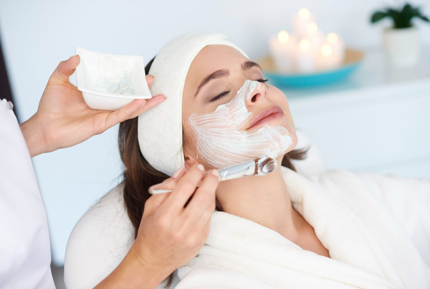 Woman getting facial mask applied at a spa; relaxed, white robe, candles in background.