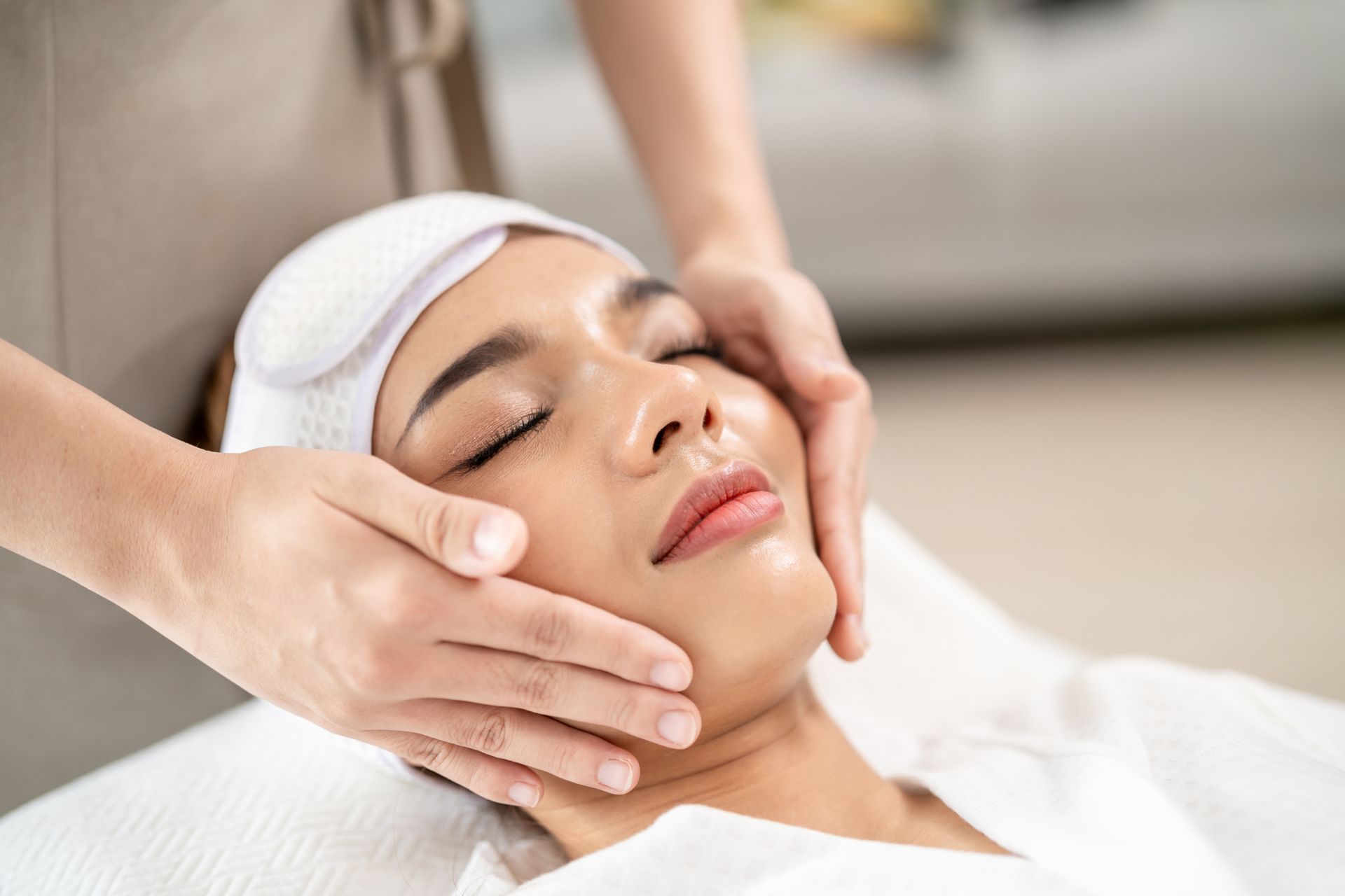 A woman with closed eyes is lying down, receiving a gentle facial massage. 