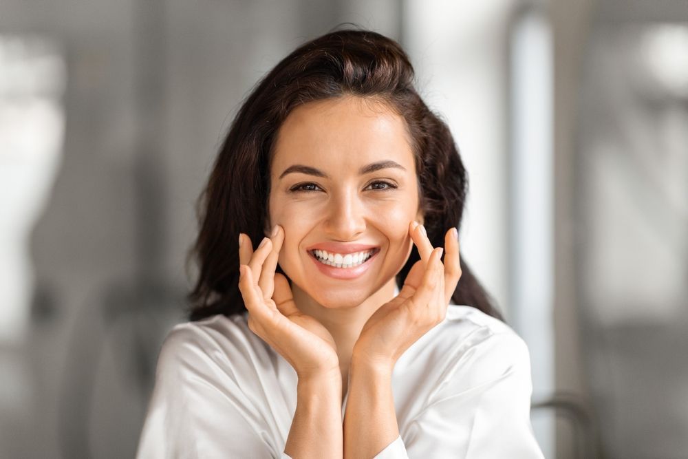 Woman with dark hair smiles, touching her cheeks, white top, blurred background.