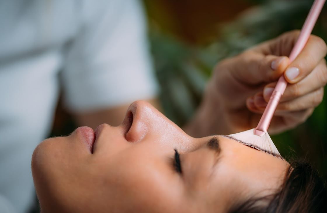 woman lying down with her eyes closed while a cosmetic brush is used to apply a product