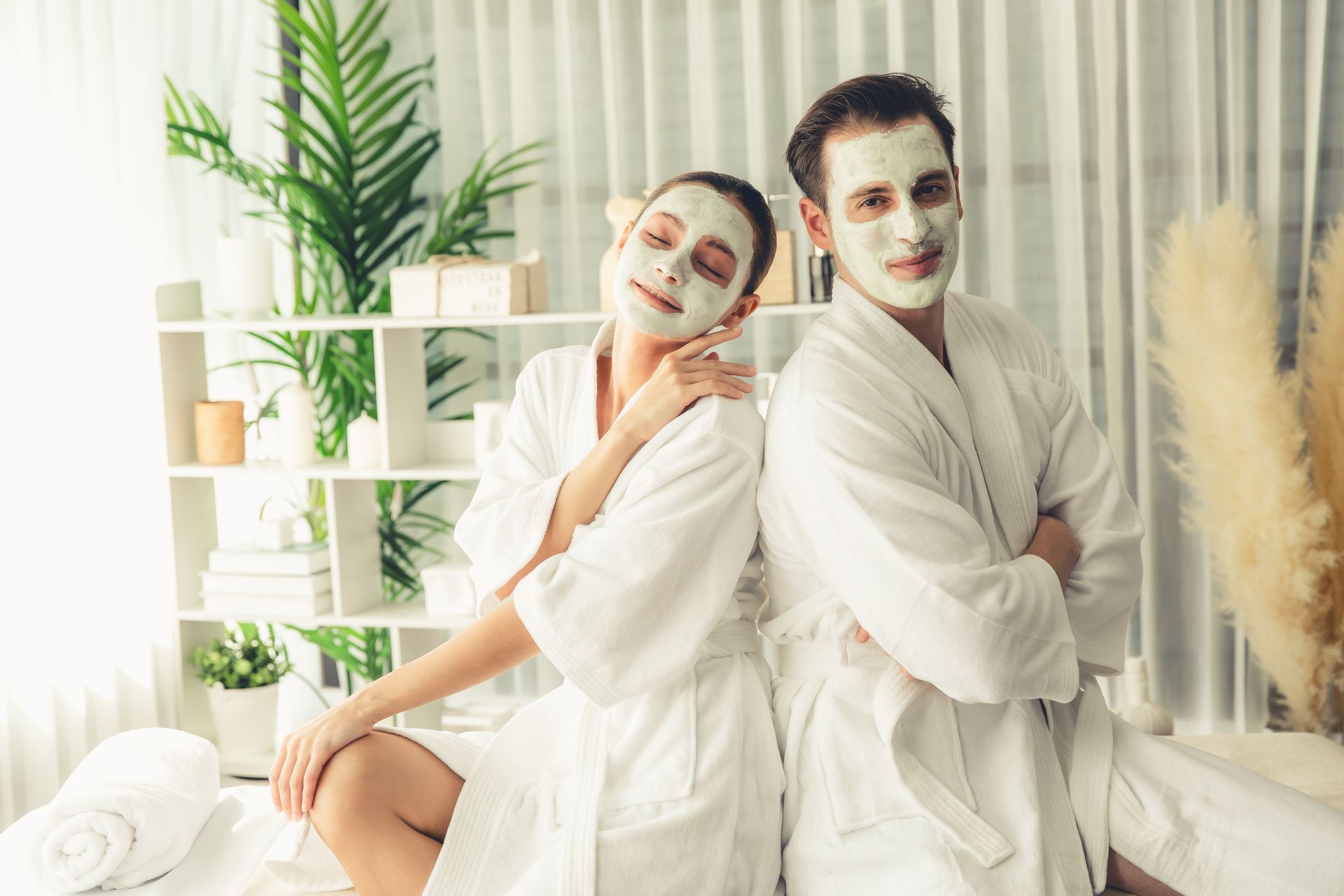 A couple in white robes and face masks relaxing at a spa, leaning back-to-back. The woman smiles, while the man has arms crossed.