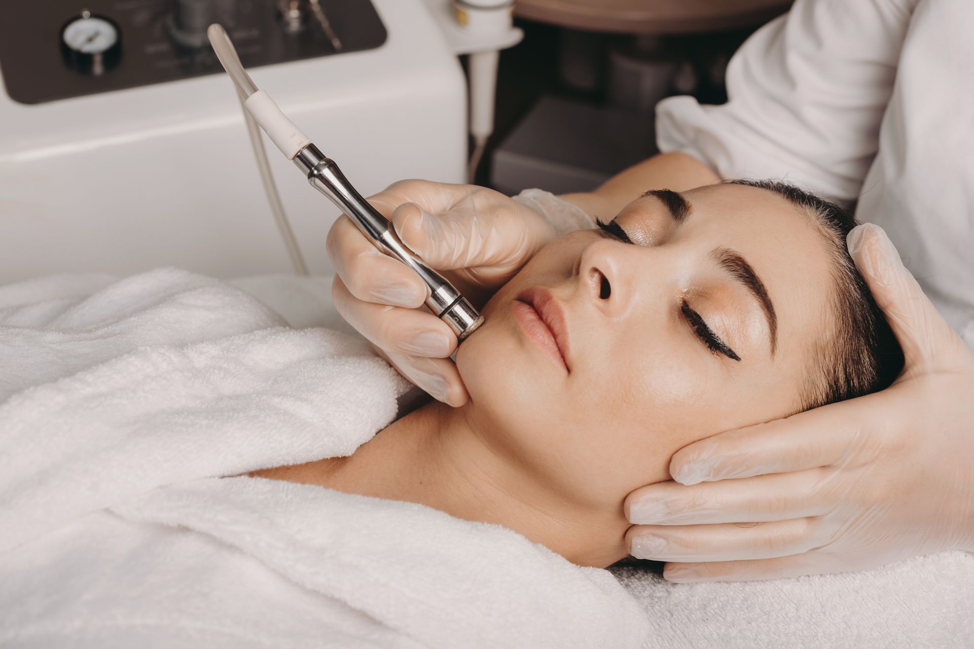Woman receiving a diamond microdermabrasion treatment at a spa, held by gloved hands.