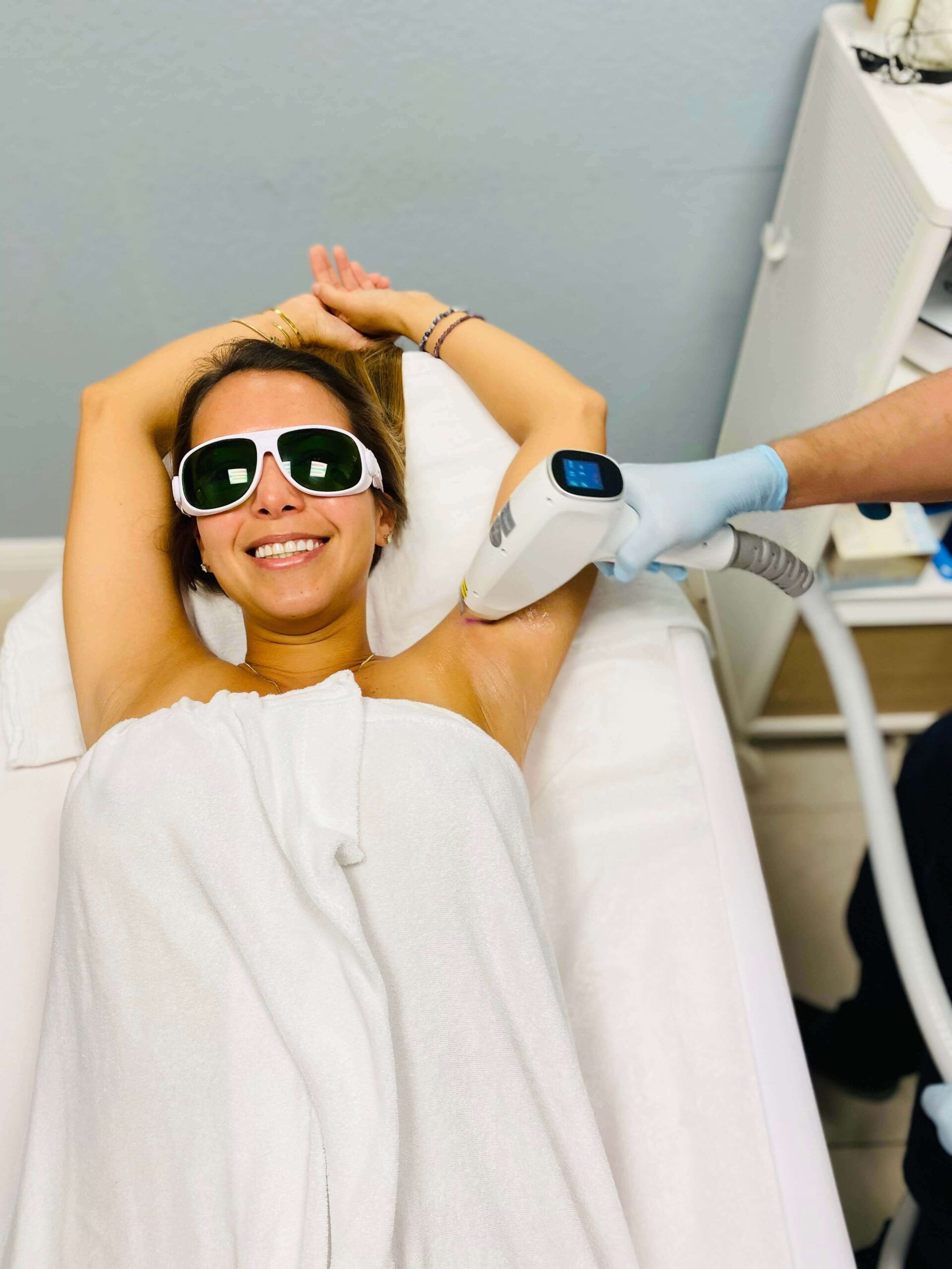 A woman wearing sunglasses is laying on a bed getting a laser hair removal treatment.