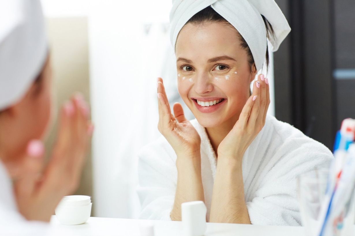 A woman with a towel wrapped around her head is applying cream to her face in front of a mirror.