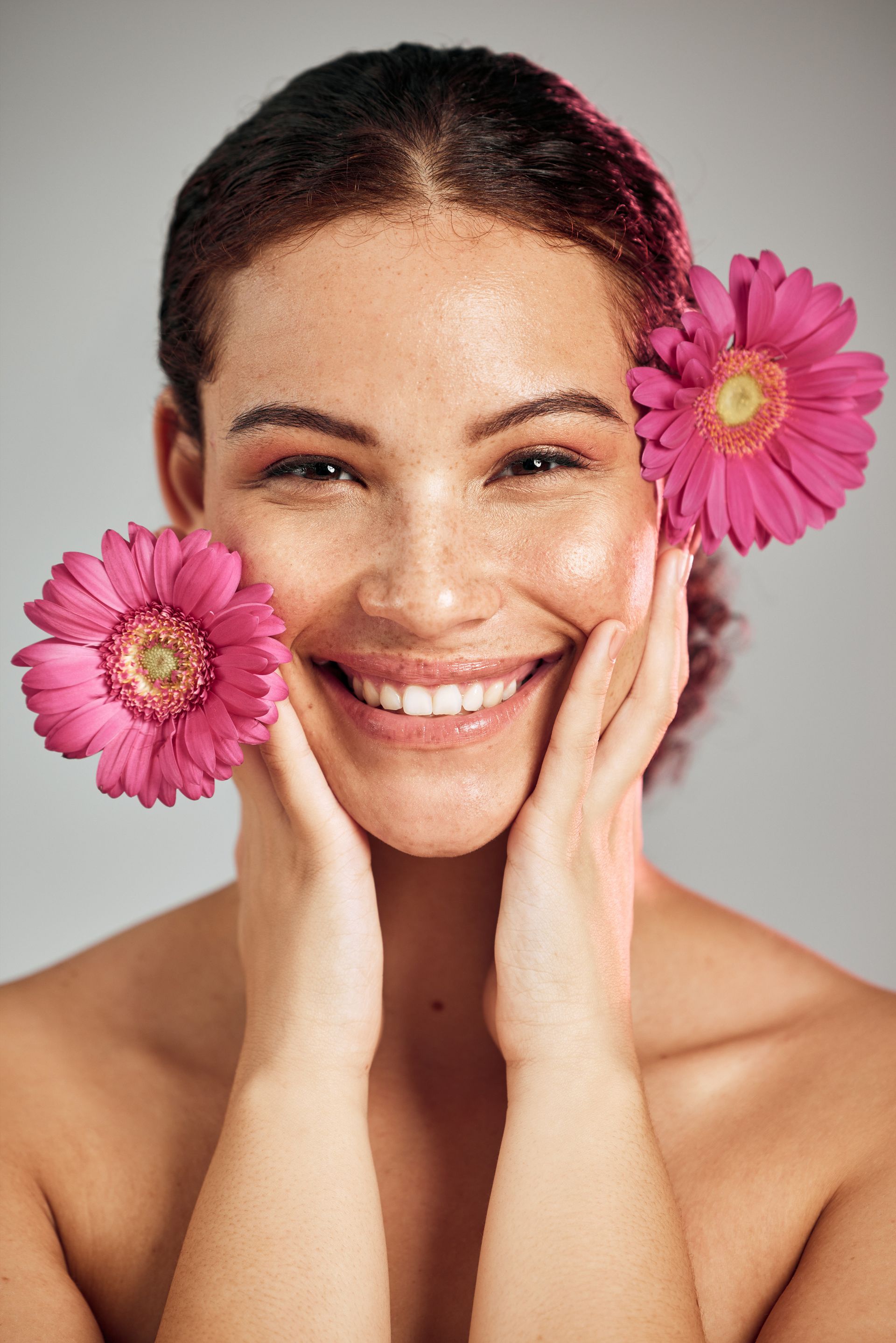 A woman is smiling with two pink flowers in her hair.