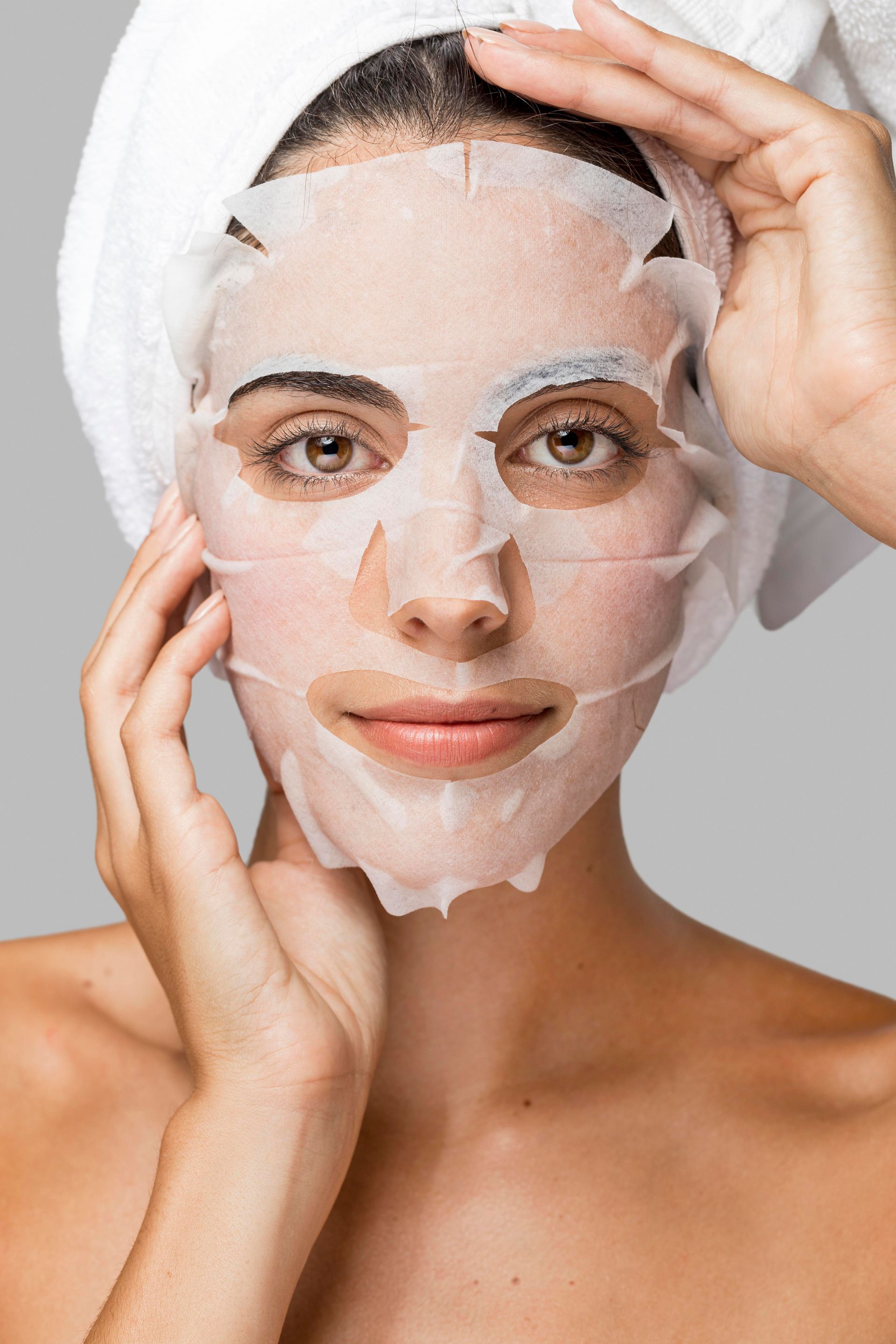 A woman with a towel wrapped around her head is wearing a sheet mask on her face.