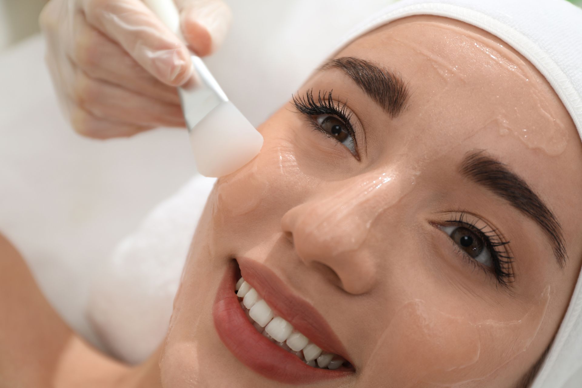 Woman receiving a facial treatment, with a technician applying a product to her cheek with a brush. She smiles.