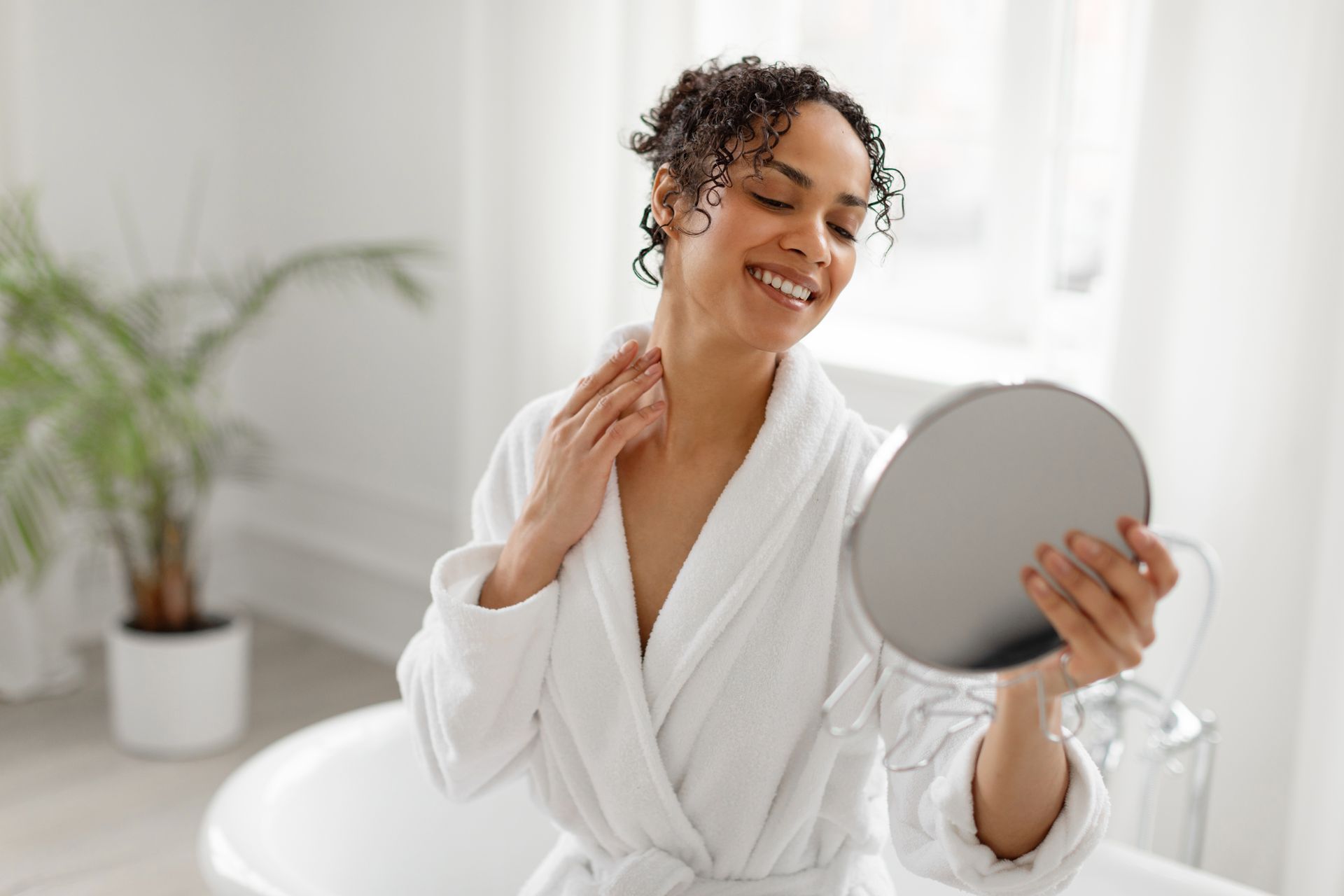 Woman in white robe looking at herself in a hand mirror in a bright bathroom.
