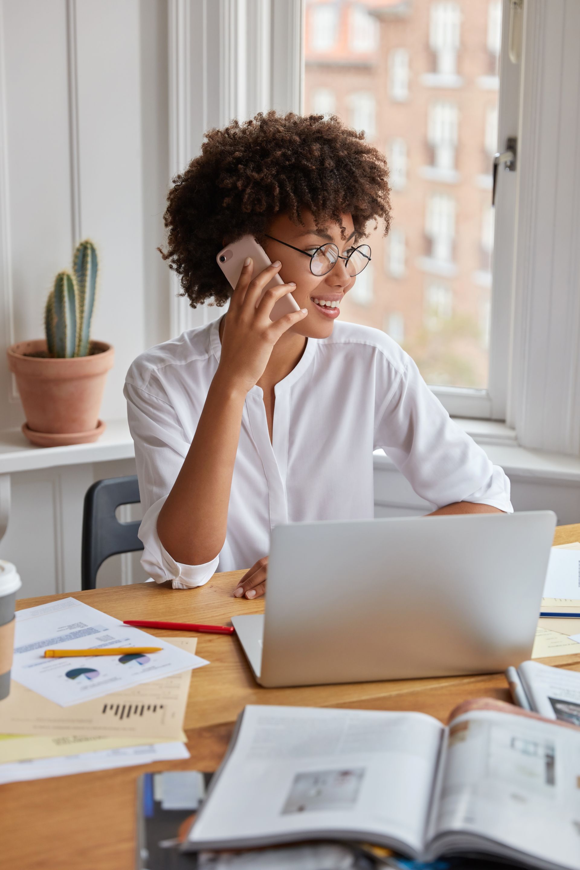 A woman is sitting at a desk talking on a cell phone while using a laptop computer.