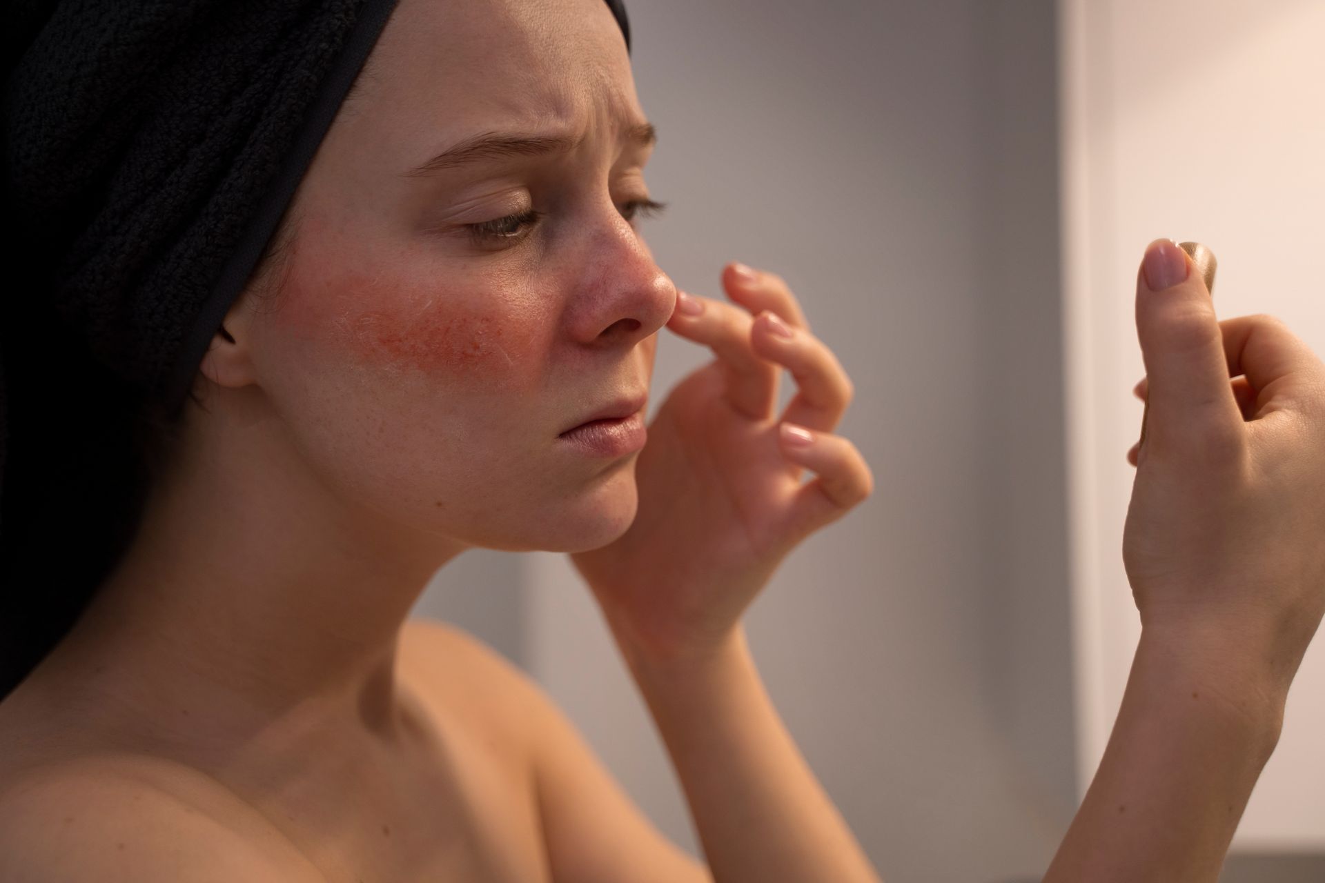Woman with a red, inflamed face looking at her reflection, touching her nose. Dark hair wrap, neutral background.