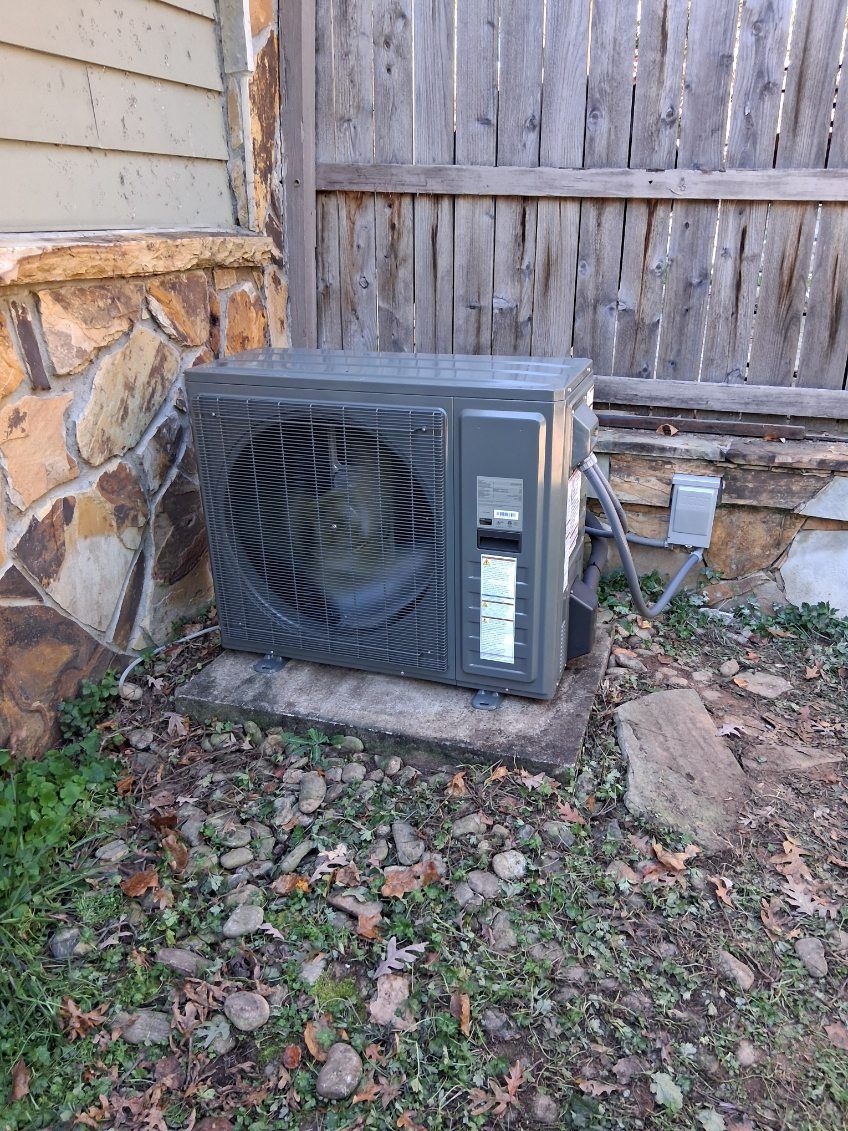 Air conditioning unit against a weathered wall and wooden fence, set on a concrete slab amidst foliage.