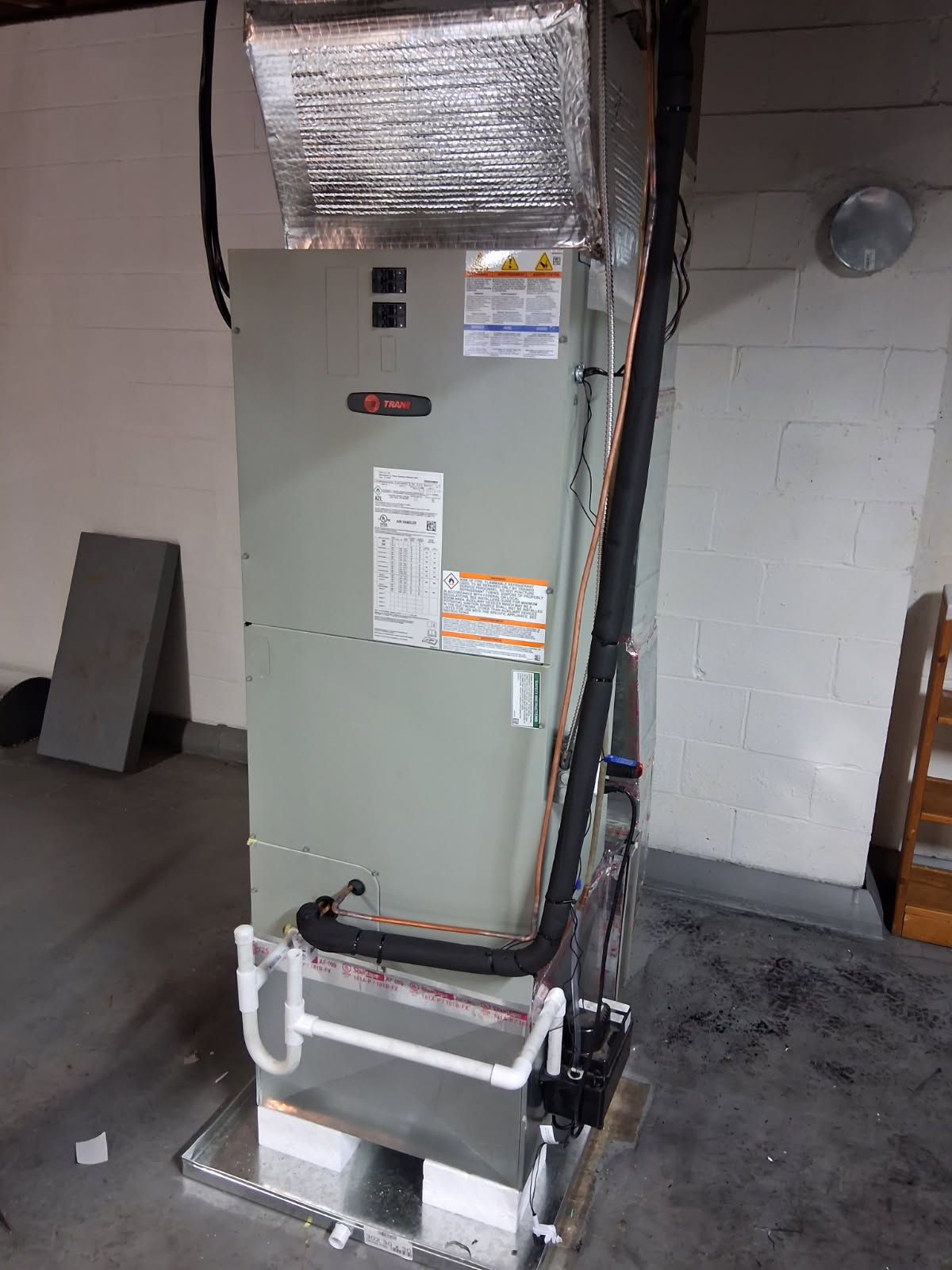 An HVAC unit stands in a basement against a white wall. Black insulated lines run along the unit.
