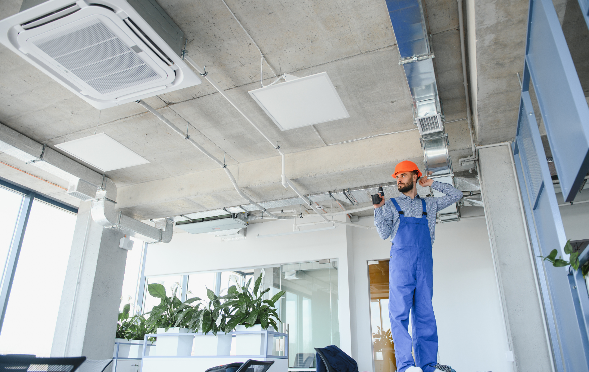 A person in overalls and a hard hat inspecting ceiling ventilation in an office.