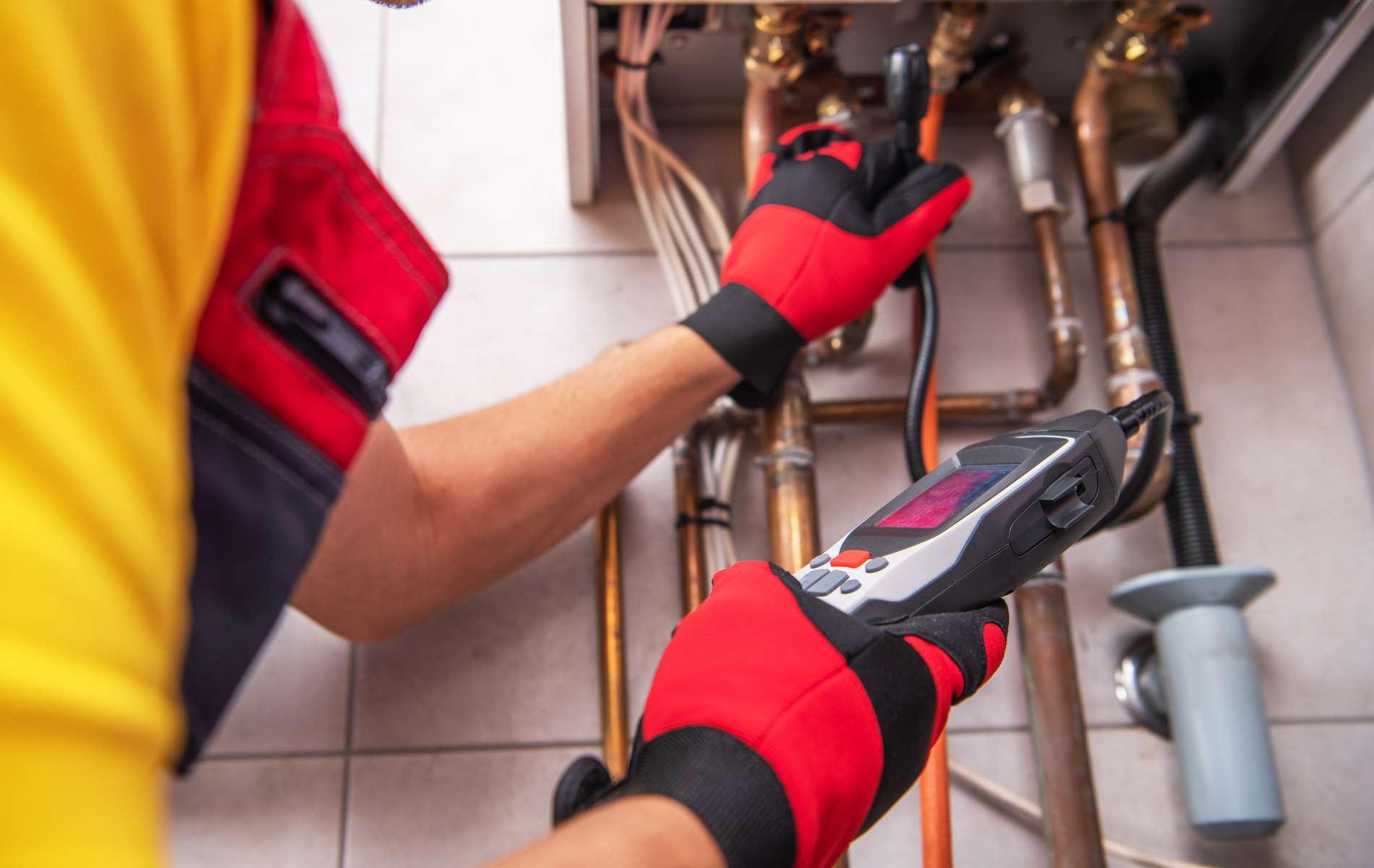 A person in red work gloves inspecting pipes with a device near a boiler.