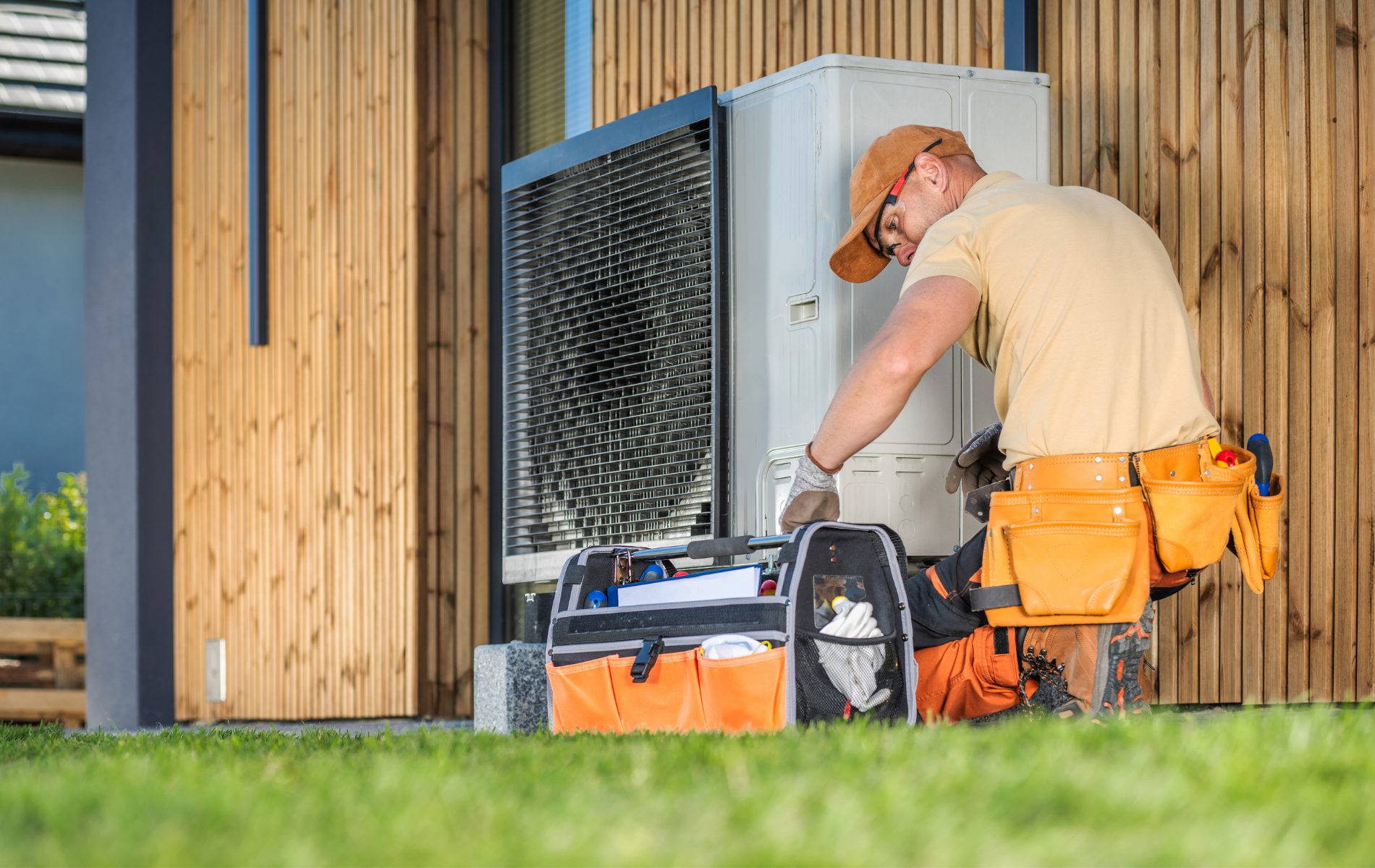 A worker in orange tool belt fixes an HVAC unit outside a wooden building on green grass.