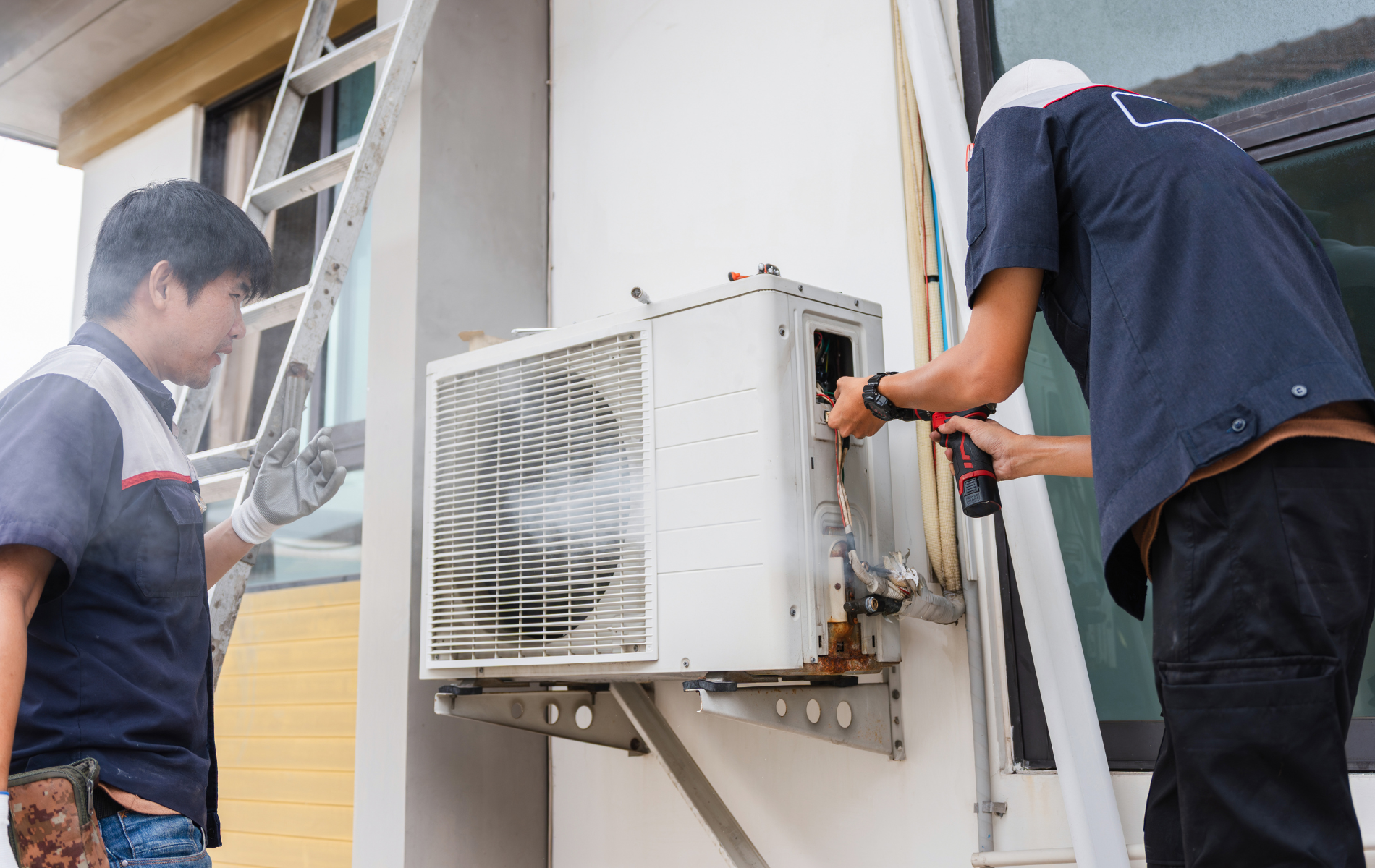 Two HVAC technicians repair an outdoor air conditioning unit mounted on a wall.