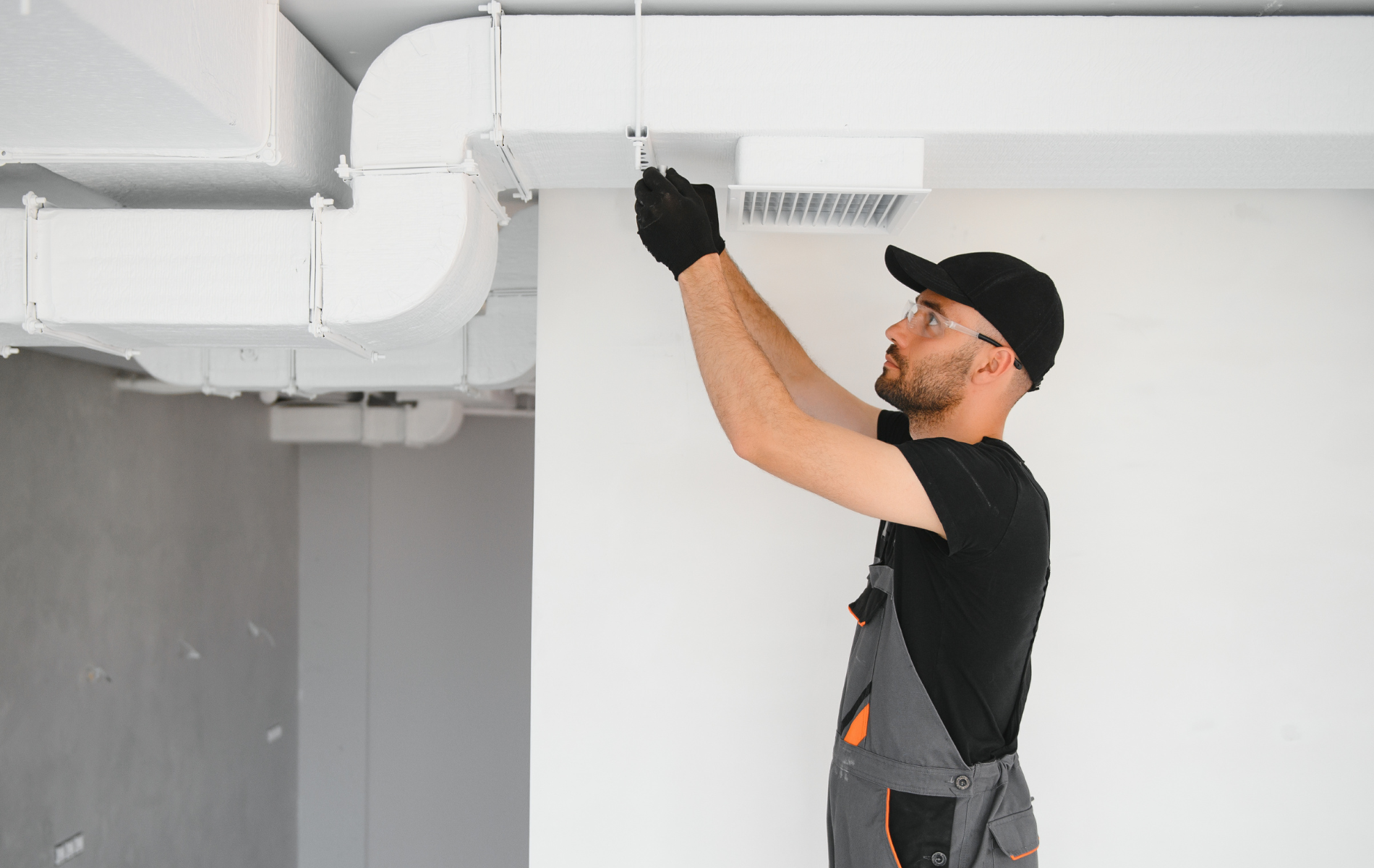 HVAC technician installing ventilation ductwork on a white wall; wearing black gloves and hat.