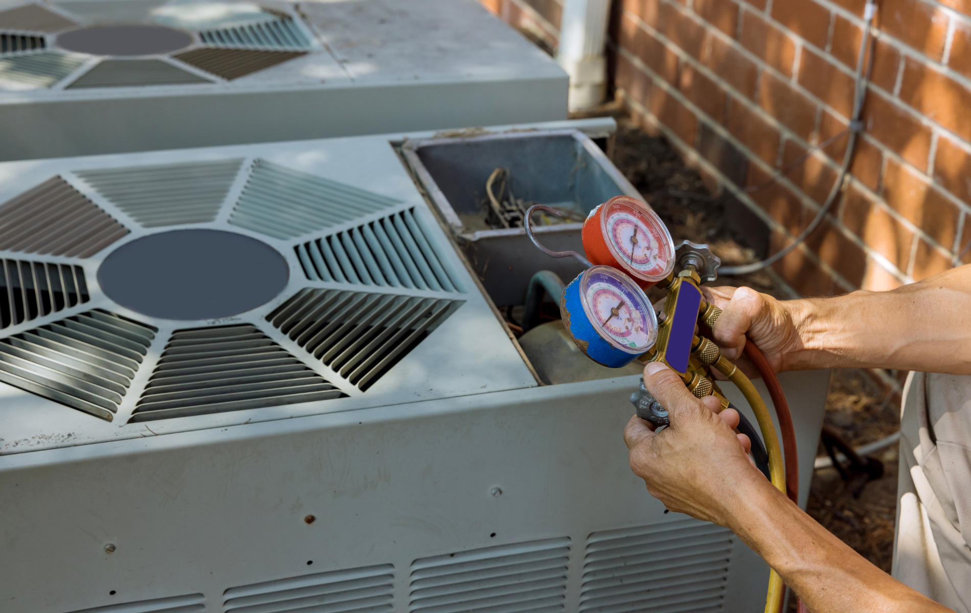 HVAC technician using gauges to service an air conditioning unit. Outdoors, near a brick wall.