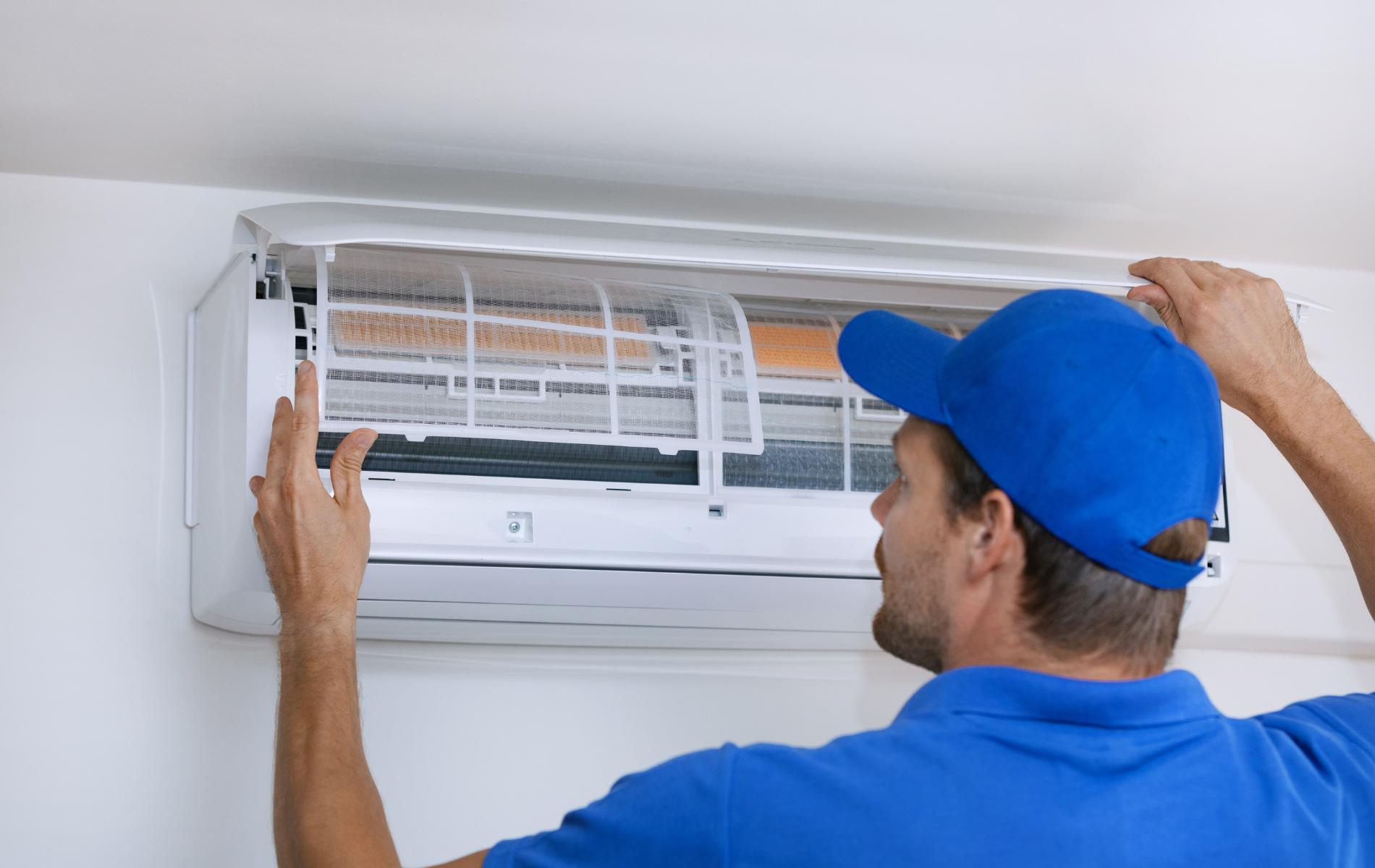 Person in blue cap and shirt replacing an air conditioner filter on a white wall.